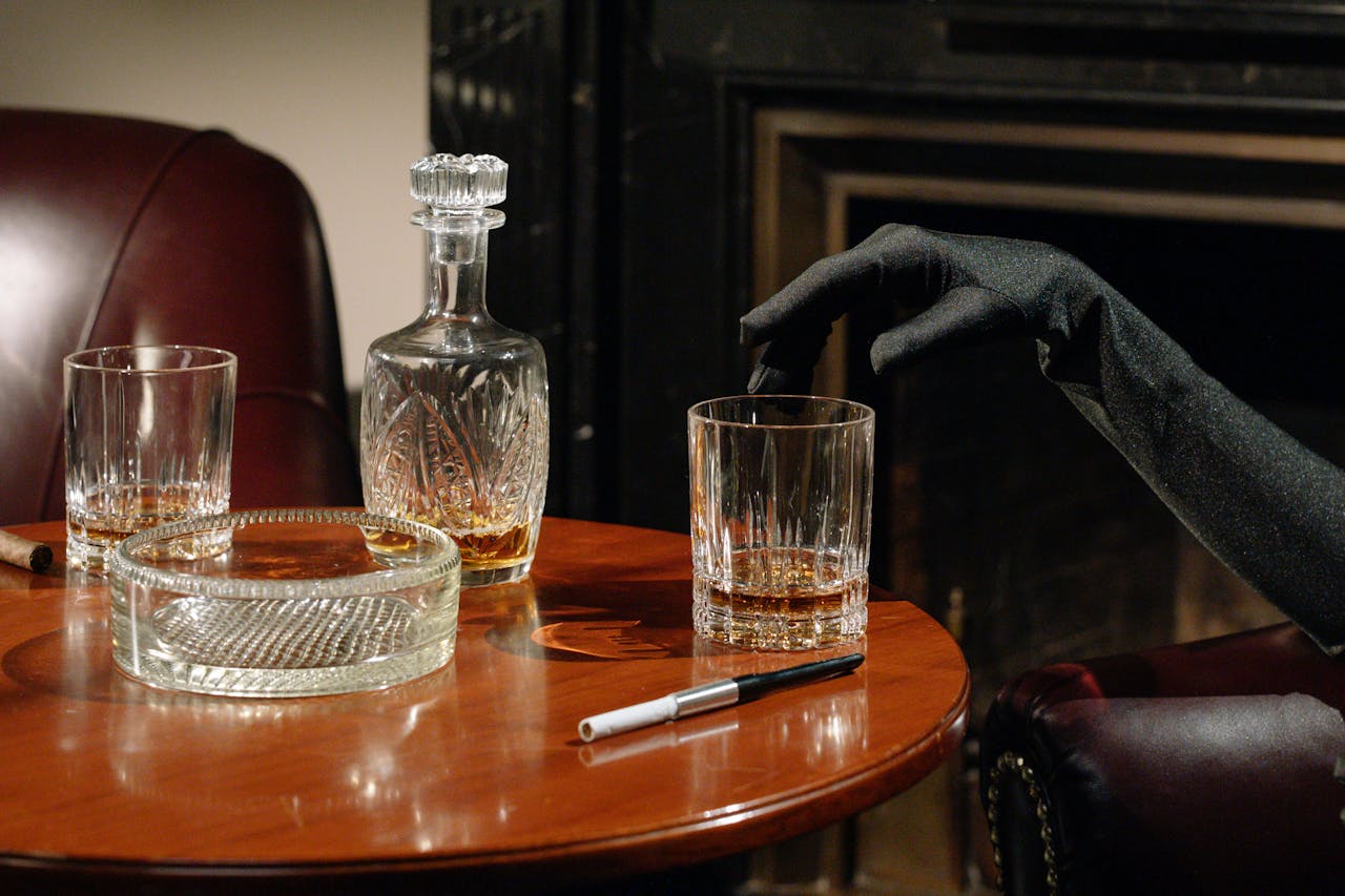 A sophisticated setup with whiskey, cigar, and glove on a wooden table by the fireplace.