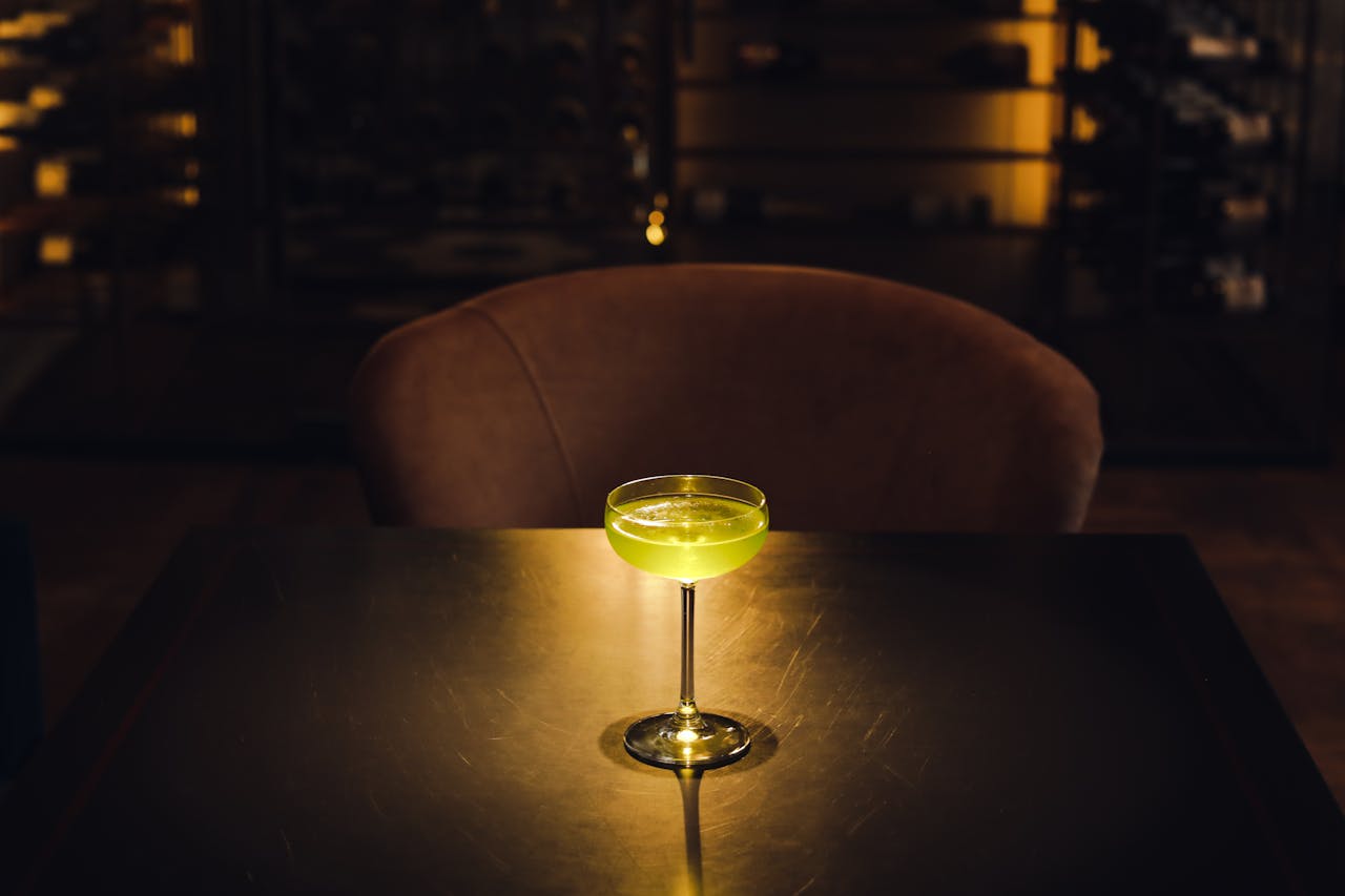 A stylish green cocktail in a glass sits on a dimly lit bar table, creating a moody atmosphere.